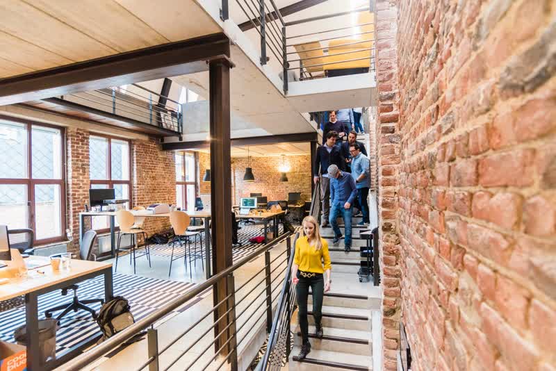 People ascend and descend a staircase in a contemporary open-plan office with exposed brick walls and wooden ceilings.